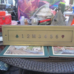 May include: A vintage brass elevator panel with numbers 1 through 6, an up arrow, and a down arrow. The panel is rectangular and has a slightly aged appearance. The numbers and arrows are in a simple, clear font.