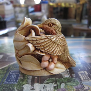 May include: A ceramic figurine of a hummingbird perched on a flower. The hummingbird is brown and beige with a red breast. The flower is pink and white.