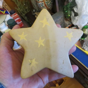 May include: A beige ceramic star with five points, decorated with five yellow stars. The star is slightly textured and has a worn look.