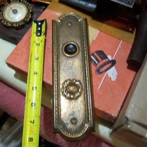 May include: An antique brass door plate with a central button and decorative details. A yellow tape measure shows the door plate is approximately 8 inches long. A small illustration of a top hat and gloves is visible.