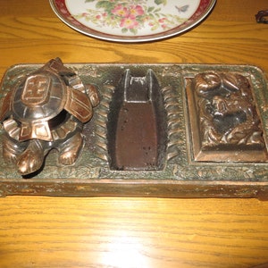 May include: A vintage metal turtle ashtray with a rectangular shape and a green patina. The ashtray has three compartments, one for cigarettes, one for matches, and one for a turtle figurine.