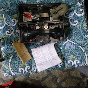May include: An open, olive-green vintage field telephone with a black handset and various internal components. Red and black cables, a green strap, and paperwork are visible. The phone rests on a patterned blue and green fabric.