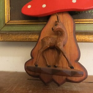 May include: A wooden wall decor featuring a carved deer standing under a red mushroom with white polka dots. The mushroom is attached to a wooden plaque with a carved tree trunk design.
