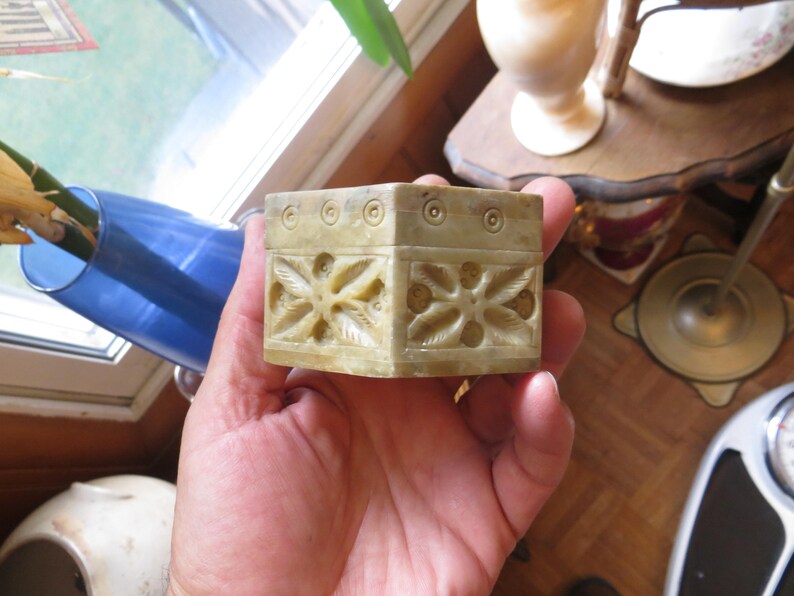May include: A small, square, carved stone box with intricate floral designs. The box is made of a light-colored stone, possibly marble or alabaster.
