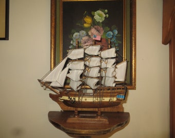 May include: A wooden model ship with white sails and an American flag. The ship is on a wooden shelf in front of a framed painting of flowers.