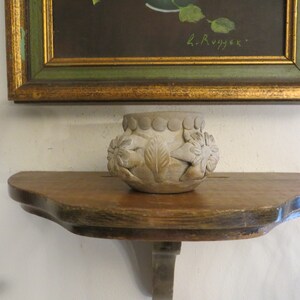May include: A small, white ceramic pot with a decorative design of flowers and leaves sits on a wooden shelf. The pot is about 4 inches in diameter and 3 inches tall.