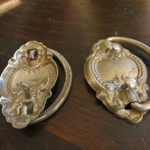 May include: Two antique silver-toned drawer pulls with ornate designs. One pull has a ring handle, while the other has a flat plate with a screw hole.