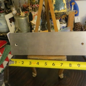 May include: A rectangular, brushed metal plate with two drilled holes, approximately 10 inches long. A yellow measuring tape is placed below the plate, showing the length. The plate is displayed on a small wooden easel.