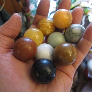 May include: A hand holding a collection of nine smooth, round, polished stones in various colors, including brown, yellow, white, black, and green.