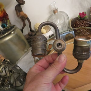 May include: A brass lamp socket with two sockets and a curved arm. The sockets are both threaded for standard light bulbs.