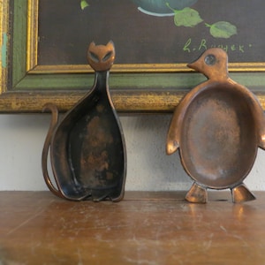 May include: Two vintage copper trinket dishes shaped like a cat and a penguin. The cat is black and the penguin is brown. The dishes are sitting on a wooden surface.