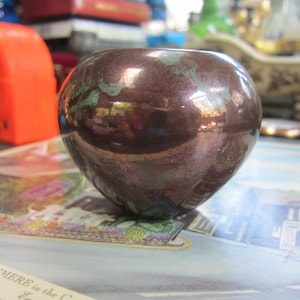 May include: A small, round, brown ceramic bowl with a metallic sheen and green flecks. The bowl is sitting on a surface with a printed design.