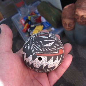 May include: A small, round, black and white pottery ornament with a red and blue design. The ornament has a small hole in the center.