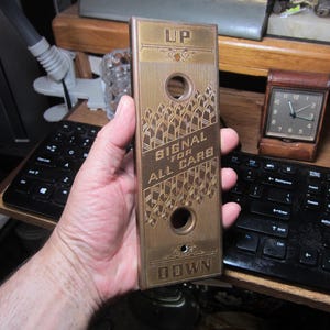 May include: A vintage brass elevator call button with the words "UP" and "DOWN" at the top and bottom. The center reads "SIGNAL FOR ALL CARS" in a decorative font. Two circular holes are visible.