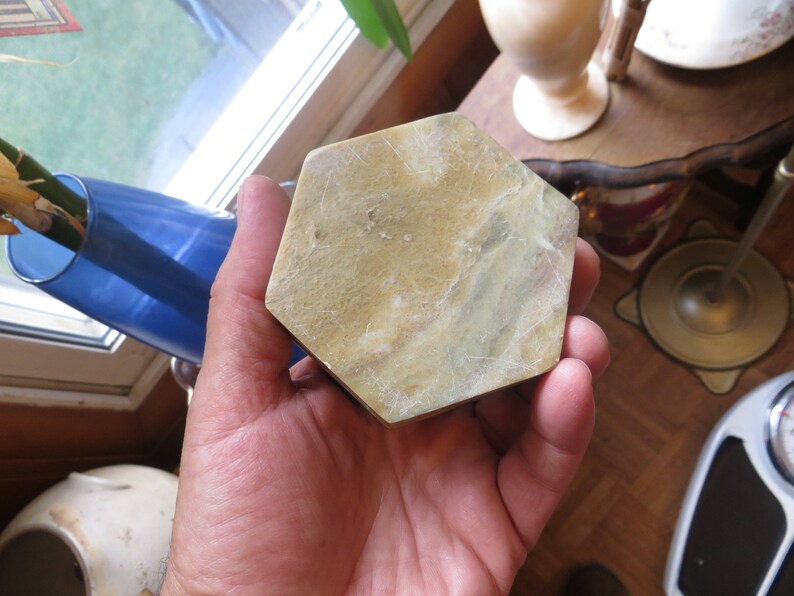 May include: A hand holding a hexagonal shaped, light green and brown stone coaster.