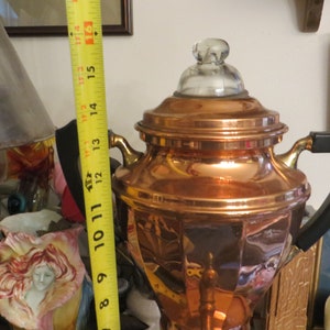 May include: A copper percolator coffee pot with a glass lid and a black handle. The pot is sitting on a table with a measuring tape in the background.