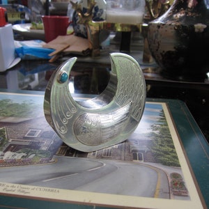 May include: A silver-colored, bird-shaped decorative object with intricate engraved details. It features a turquoise-colored stone at the top and is likely a decorative item or a unique piece of art. The object is resting on a placemat with a scenic image.