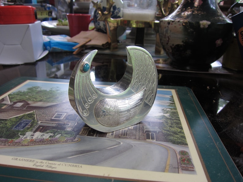 May include: A silver-colored, bird-shaped decorative object with intricate engraved details. It features a turquoise-colored stone at the top and is likely a decorative item or a unique piece of art. The object is resting on a placemat with a scenic image.