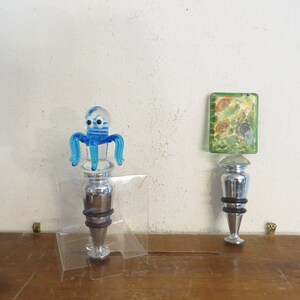 May include: Two decorative bottle stoppers. One is silver with a blue glass octopus on top. The other is silver with a green and white glass square on top.