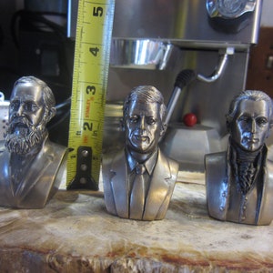 May include: Three silver-toned bust sculptures of historical figures. The sculptures are on a wooden surface. The bust on the left has a beard and is wearing a suit. The bust in the middle is wearing a suit and tie. The bust on the right is wearing a white shirt with a dark vest.