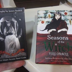 May include: Two tarot card decks. The first deck is black with a silver image of a woman in a white dress holding a harp. The text on the box reads "Salem Arcanum Tot 22 Major Arcana Tarot Cards". The second deck is red with a green image of a woman in a red dress with a crown on her head. The text on the box reads "Seasons of the Witch Yule Oracle Lorriane Anderson & Juliet Diaz Illustrated by Giada Rose".