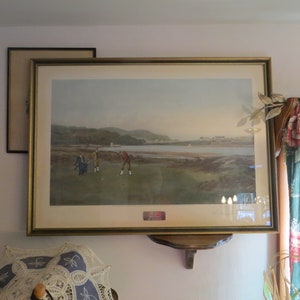 May include: A framed print of a golf scene with three golfers on a green course. The scene is set against a backdrop of a body of water and hills. The print is in a gold frame and is hanging on a wall.