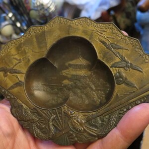 May include: A brass trinket dish shaped like a fan with three small bowls. The bowls are decorated with a pagoda and trees. The fan is decorated with birds and flowers.
