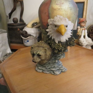 May include: A ceramic figurine of a bald eagle with a brown bear cub perched on its back. The eagle has a yellow beak and white head feathers. The bear cub is brown and has a black nose. The figurine is set on a rocky base.