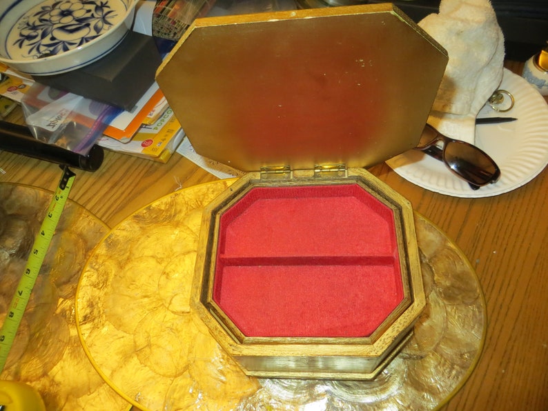 May include: A gold octagonal wooden jewellery box with a red velvet interior. The box has a hinged lid and two compartments.