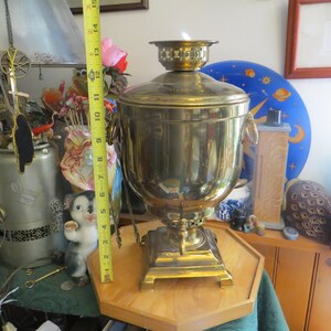 May include: A gold-colored samovar, a traditional Russian tea kettle, with a round body and a spout. The samovar is sitting on a wooden stand with a hexagonal shape.