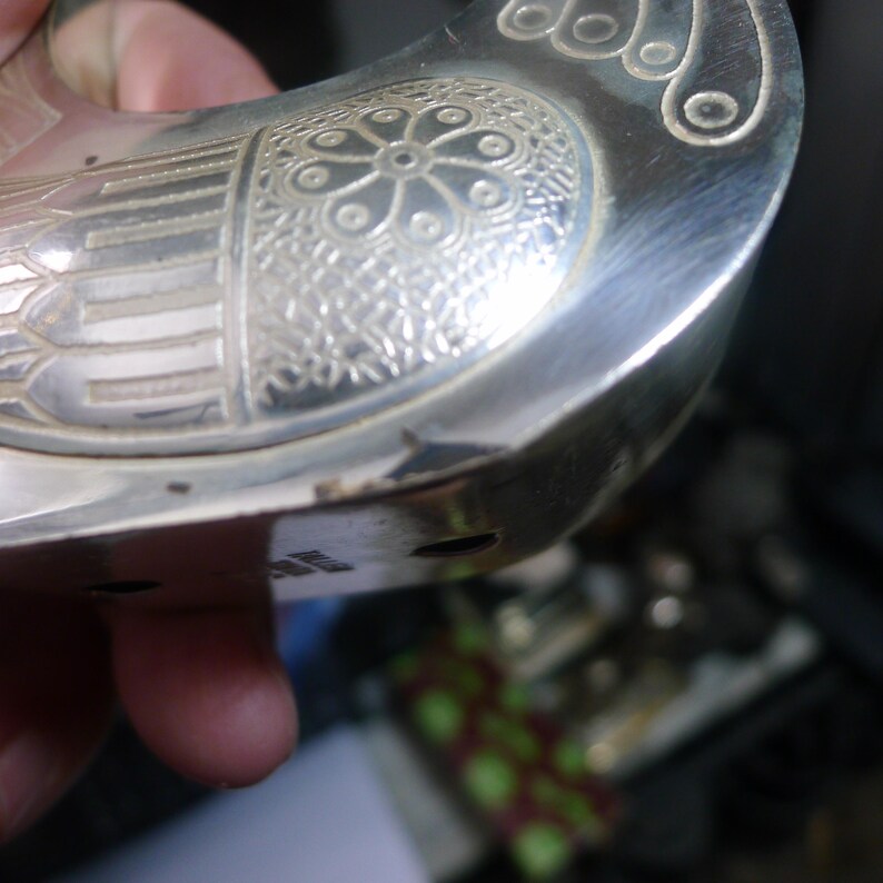 May include: Close-up of a silver-colored metal object with intricate floral and geometric patterns. The object has a curved shape with a handle-like extension and decorative detailing. The surface is polished and reflective.