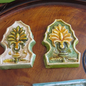 May include: Two ceramic tiles with a green, yellow, and white floral design. The tiles are shaped like a pointed arch and have a raised design. The tiles are on a wooden surface.