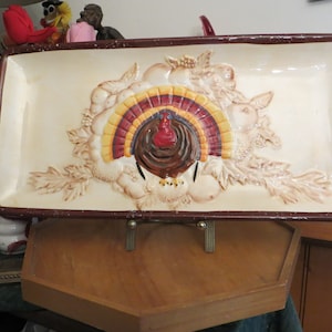 May include: A ceramic serving tray with a raised design of a turkey with brown, red, and yellow feathers. The tray has a brown rim and is on a wooden stand.