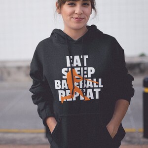 May include: A black hoodie with a white graphic that says "Eat Sleep Baseball Repeat" with an orange baseball player silhouette. The hoodie has a drawstring hood and a kangaroo pocket.