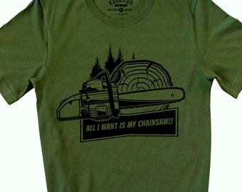 Op de afbeelding: Een olijfgroen T-shirt met korte mouwen en een zwarte afbeelding van een kettingzaag, een boomstronk en dennenbomen. Onder de afbeelding staat in een rechthoekig kader de tekst "ALL I WANT IS MY CHAINSAW!!". Het label in de nek vermeldt "KEBEKER".