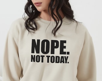 以下が含まれることがあります： 「NOPE. NOT TODAY.」のテキストが黒い太字でプリントされたベージュのスウェットシャツ。スウェットシャツは長袖でクルーネックです。モデルはデニムショーツを着用しています。