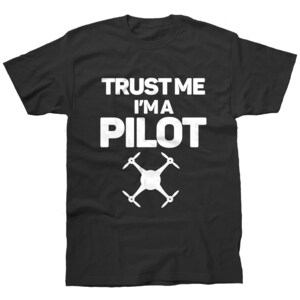 May include: A black short-sleeved t-shirt featuring the white text "TRUST ME I'M A PILOT" printed in a bold, sans-serif font. Below the text is a white graphic illustration of a four-propeller drone viewed from directly above.