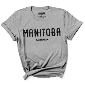 May include: A heather gray t-shirt with the word "MANITOBA" in black, arched text, and "CANADA" below it. The shirt has rolled-up sleeves and a crew neck. The brand name "XEREKER DESIGN" is printed at the top.