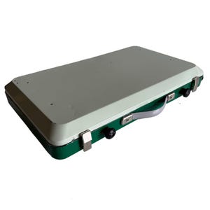 Peut inclure: Un réchaud de camping portable rectangulaire plié en mallette fermée. L'extérieur présente un panneau supérieur vert clair et une base vert foncé, fixés par des loquets en métal argenté. Une poignée en plastique gris est fixée sur le côté.