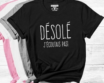 Op de afbeelding: Een zwart t-shirt met korte mouwen en de witte tekst DÉSOLÉ J'ÉCOUTAIS PAS! op de voorzijde. Het t-shirt wordt getoond met een stapel andere t-shirts in roze, witte en grijze kleuren die onder het zwarte kledingstuk zichtbaar zijn.