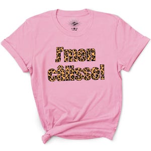 Peut inclure: Un t-shirt rose avec l'inscription "J'men câlisse!" en imprimé léopard. Le t-shirt a un col rond et des manches courtes. Le texte est en gras, en police sans-serif.