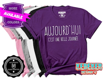 Op de afbeelding: Een paars t-shirt met korte mouwen en witte tekst die luidt AUJOURD'HUI C'EST UNE BELLE JOURNÉE. Het t-shirt wordt getoond in een stapel met andere t-shirts in roze, grijs, wit en zwart, allemaal met hetzelfde witte bedrukte tekstontwerp.