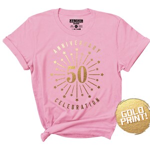 May include: Pink t-shirt with gold foil print that says "Anniversary Celebration" with a firework design and the number 50 in the center.