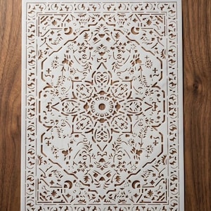 May include: A white, rectangular stencil with a detailed, symmetrical floral and geometric design. The stencil features cut-out patterns of flowers, leaves, and other shapes, creating a decorative border and central medallion. The stencil is placed on a wooden surface.