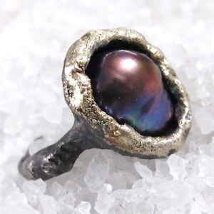 May include: A unique ring featuring a dark, iridescent pearl set in a textured, gold-toned bezel. The ring's band is a dark, oxidized metal, creating a striking contrast. The pearl displays hues of purple, blue, and green, adding depth and visual interest to the jewelry.