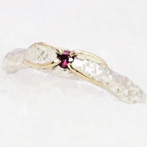 Op de afbeelding: Een zilveren ring met een getextureerde afwerking en een kleine rode edelsteen in het midden. De ring is versierd met een dunne gouden band.