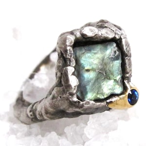 May include: A silver ring with a square-shaped green gemstone set in a textured silver band. A small blue gemstone is set in a gold accent on the side of the ring.