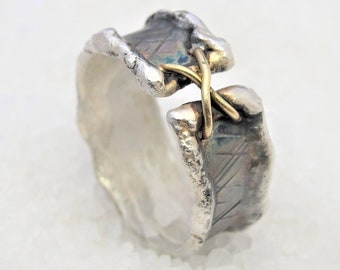 May include: A silver ring with a textured finish and a gold band that crosses over the top. The ring is designed with a unique, asymmetrical shape.