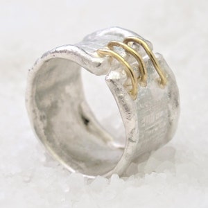Könnte beinhalten: Ein silberner Ring mit einer strukturierten Band und drei goldenen Akzenten. Der Ring ist in einer einzigartigen, asymmetrischen Form gestaltet.