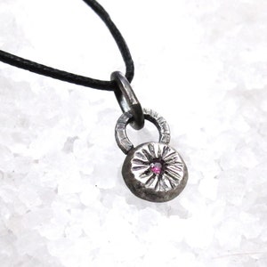 May include: A silver pendant necklace with a small pink gemstone in the center of a textured circular design. The pendant is attached to a black cord.